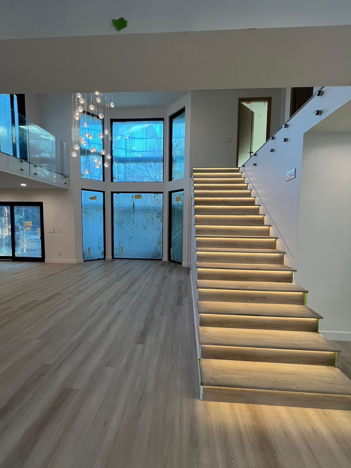 Open interior with illuminated wood-look stairs, glass railings and tall windows during finishing work